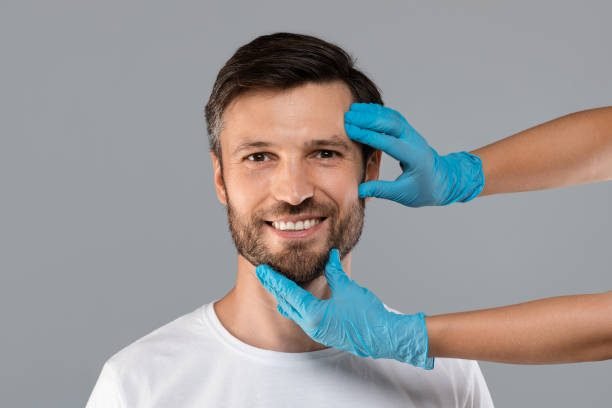 Cosmetician hands in protective medical gloves touching smiling man face over grey studio background. Plastic surgery and aesthetic cosmetology concept. Cheerful handsome man ready for plastic surgery